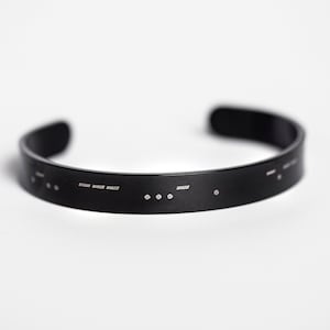 May include: A black cuff bracelet with Morse code detailing. The bracelet is made of a dark, matte material and features a series of dots and dashes. The open-ended design allows for easy wear. The bracelet is displayed on a white background.