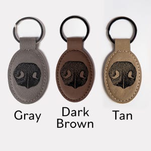May include: Three oval-shaped keychains in gray, dark brown, and tan leather-like material. Each features a black engraved dog nose print in the center and a black metal split ring at the top. The text Gray, Dark Brown, and Tan is visible.