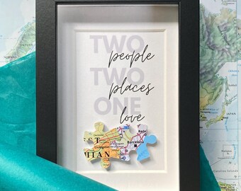 May include: A framed print with the text "Two people, two places, one love" and a map puzzle of two locations. The puzzle pieces are joined together in the center of the frame.