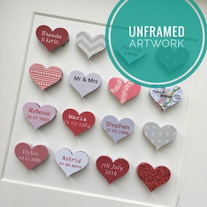 May include: A white frame with a light gray background containing 12 layered paper hearts with different patterns and colors. Each heart has a name and date printed on it. The text "UNFRAMED ARTWORK" is printed in black on a teal circle at the top right corner of the frame.