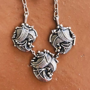 May include: A silver necklace with three pendant charms, each featuring a cluster of stylized leaves. The leaves are textured and have a slightly raised design.