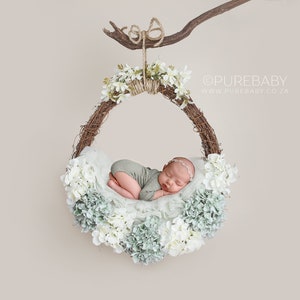May include: A newborn baby sleeping in a white and green flower wreath hanging from a branch. The wreath is made of twigs and has white and green flowers around it.