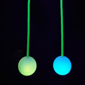 Op de afbeelding: Twee gloeiende poi-ballen, één groen en één blauw, hangen aan felgroene koorden tegen een zwarte achtergrond. De ballen worden vastgehouden door handen die worden verlicht met paars licht. De poi-ballen zijn een levendig en opvallend rekwisiet.
