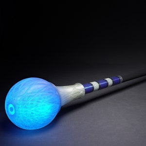 Puede incluir: Un bastón brillante azul y blanco con un orbe redondo en cada extremo. El orbe en el extremo del bastón está iluminado con una luz azul brillante.