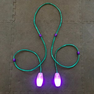 Puede incluir: Dos mazas de malabares luminosas de color morado con cordones verdes y cuentas moradas. Las mazas están sobre una superficie gris texturizada.