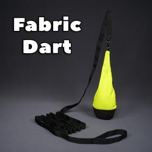 Puede incluir: Un dardo de tela amarillo con una correa negra y un mango negro. La correa está enrollada en el suelo. El texto "Fabric Dart" está escrito en letras blancas sobre un fondo negro.