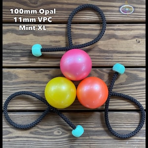 Pode incluir: Três bolas de malabarismo rosa, amarelas e laranja, cada uma presa a uma corda preta com uma pega turquesa. O texto "100mm Opal, 11mm VPC, Mint XL" é visível. As bolas estão dispostas sobre uma superfície de madeira.