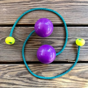 Op de afbeelding: Twee paarse ballen verbonden door een turquoise touw met gele ballen aan de uiteinden. Het touw is om de paarse ballen gewikkeld.