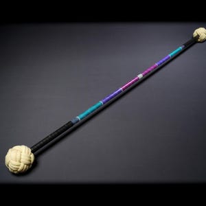 May include: A long staff featuring a black handle at each end, topped with large, cream-coloured woven rope knots. The central section of the staff is wrapped in alternating segments of teal, purple, and a small silver band in the middle.