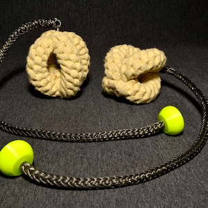 Puede incluir: Dos juguetes de cuerda beige con una cuerda trenzada negra y gris y detalles en verde brillante. Los juguetes están anudados y parecen ser para mascotas.