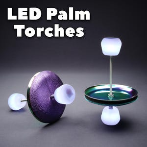 May include: Two LED palm torches with the text LED Palm Torches displayed above. One torch features a textured purple circular base, while the other has a metallic iridescent green and purple circular base. Both have white glowing LED light caps.