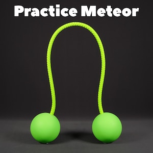 May include: A neon green practice meteor toy with two green spheres connected by a bright green rope. The text "Practice Meteor" is displayed in white at the top of the image. The toy is set against a dark background.