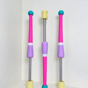 Pode incluir: Três maças de malabarismo com hastes prateadas, com pegas de espuma rosa brilhante e lavanda, extremidades esféricas azul-petróleo e tampas de tecido amarelo texturizado. As duas maças exteriores têm secções superiores cor-de-rosa.