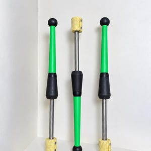 Puede incluir: Tres mazas de malabarismo con ejes metálicos, pomos negros y mangos verdes cónicos. Dos mazas presentan mechas texturizadas amarillas en la base, mientras que la maza central tiene una mecha amarilla en la parte superior y un mango verde.