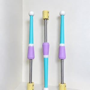 May include: Three juggling clubs with metallic shafts, featuring turquoise and lavender tapered sections, white spherical ends, and yellow textured grips at the base. The central club is inverted compared to the two outer clubs, displaying a yellow grip at the top.