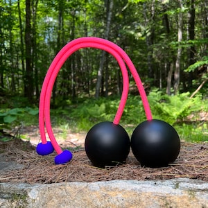 May include: Two black juggling balls connected by a vibrant pink rope. The rope curves over the balls, with blue caps at the ends. The balls are displayed on a stone surface, with a forest in the background.