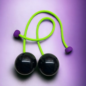 May include: A pair of black, glossy balls connected by a bright green rope with purple stoppers. The toy is set against a gradient purple background. This is a classic skill toy.