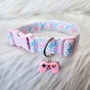 May include: A light pink dog collar with a white ribbon patterned with blue and pink video game controllers. The collar has a pink plastic buckle and a silver metal ring holding a pink controller charm.