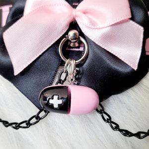 May include: A black choker with a pink satin bow and a silver charm. The charm is a pill-shaped pendant with a white cross on it.