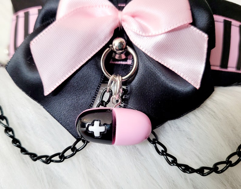May include: A black choker with a pink satin bow and a silver charm. The charm is a pill-shaped pendant with a white cross on it.