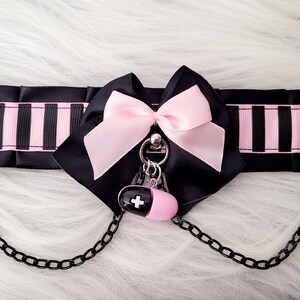 May include: Black and pink striped choker with a pink bow and a pill charm. The choker has two metal rings with chains attached.