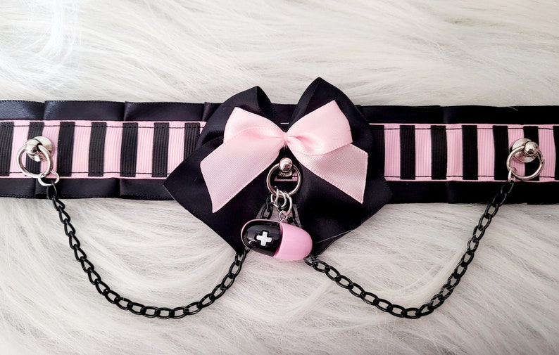 May include: Black and pink striped choker with a pink bow and a pill charm. The choker has two metal rings with chains attached.