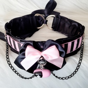 May include: Black choker necklace with pink ribbon bow and a black chain with a pink and black charm. The choker has black and white stripes.