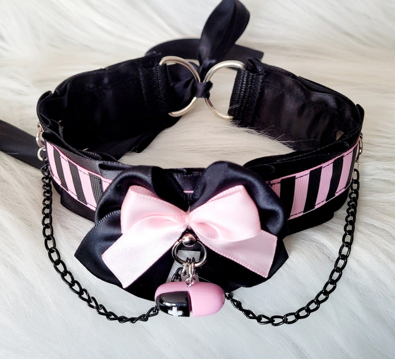 May include: Black choker necklace with pink ribbon bow and a black chain with a pink and black charm. The choker has black and white stripes.