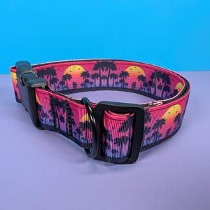May include: A pink and black dog collar with a sunset and palm tree print. The collar has a black buckle and a black D-ring.