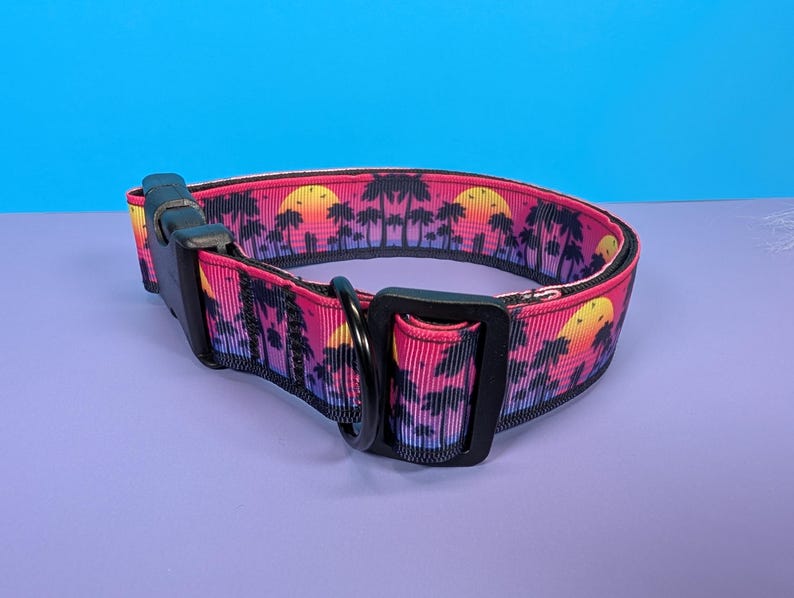 May include: A pink and black dog collar with a sunset and palm tree print. The collar has a black buckle and a black D-ring.