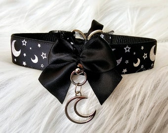 May include: Black choker necklace with a celestial design. The black ribbon features a pattern of white moons and stars. A black bow is centered on the choker, with a silver crescent moon charm hanging below.