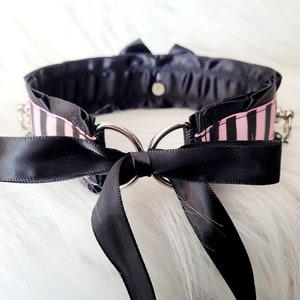 May include: A black choker with a pink and black striped ribbon and a black satin bow. The choker has two silver rings and a silver chain.