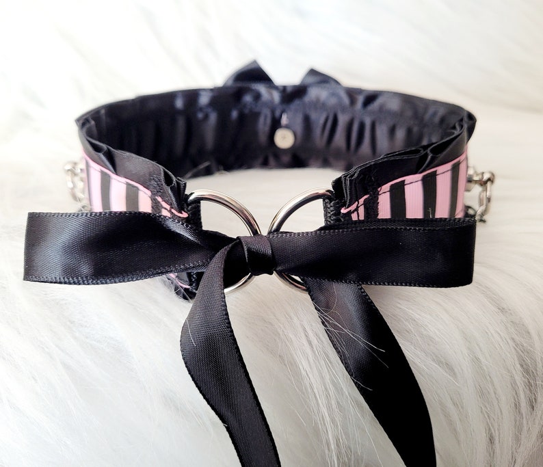 May include: A black choker with a pink and black striped ribbon and a black satin bow. The choker has two silver rings and a silver chain.