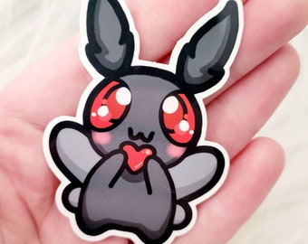 May include: A gray and black cartoon moth with large red eyes and pink cheeks holding a red heart. The moth is a sticker with a glossy finish.