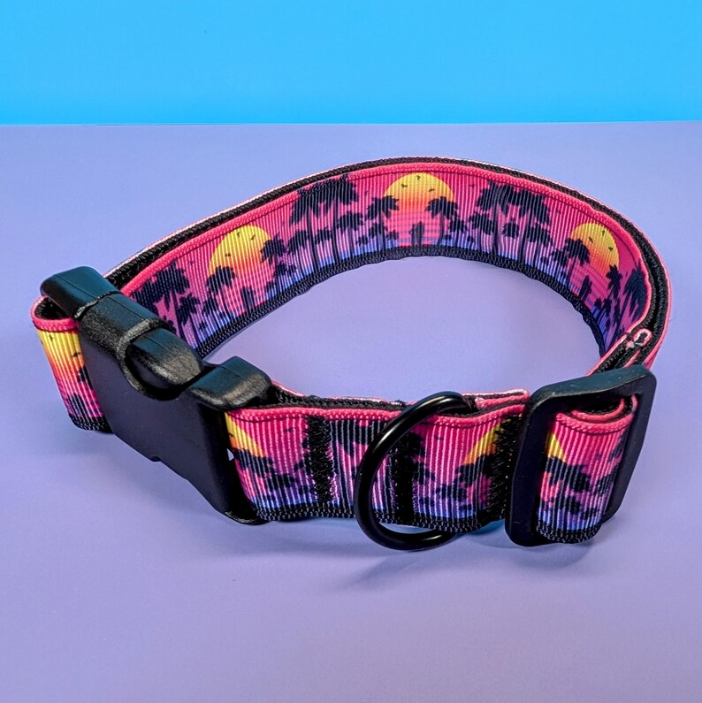 May include: A pink and purple dog collar with a sunset and palm tree print. The collar has a black buckle and a black metal D-ring.