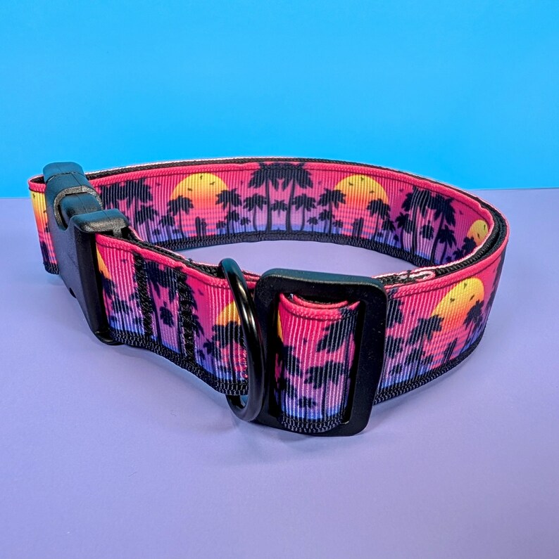 May include: A pink and purple dog collar with a black sunset and palm tree print. The collar has a black buckle and a black D-ring.