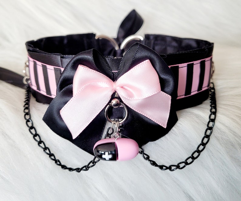 A black choker with pink and black striped ribbon and a pink bow and a large black bow. The choker has black chain attached with a pill-shaped charm.