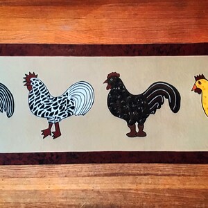 May include: A colorful painting of four roosters on a beige background. The roosters are painted in blue, black, white, and yellow. The painting is framed with a brown border.