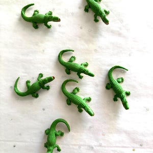 May include: Seven small, green alligator figurines are scattered on a white surface. Each alligator has a textured body, four legs, and a curved tail. The alligators have red accents on their snouts.