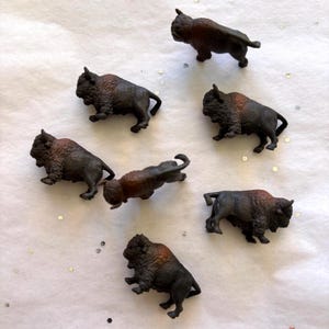 May include: A collection of small, detailed bison figurines. The figurines are primarily dark brown with lighter brown fur on their heads and shoulders. They are arranged on a white surface with scattered glitter.