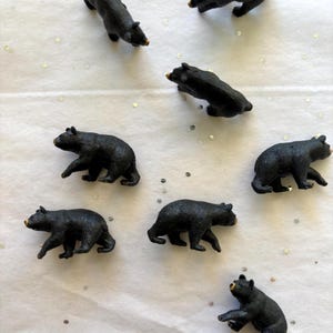 May include: A collection of small, black bear figurines with tan snouts are scattered on a white surface. The bears are in various poses, some walking and others standing. The scene is simple and playful, suitable for children's toys or decorative items.
