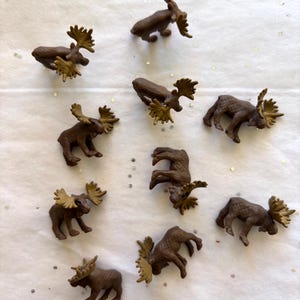 May include: A collection of small, brown moose figurines with tan antlers. The moose are in various poses, some standing and others lying down. The figurines are arranged on a white surface with scattered silver glitter.