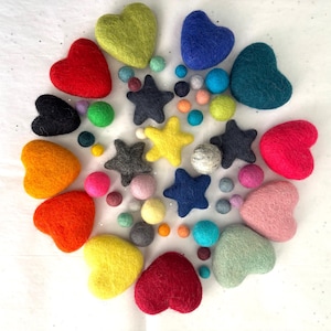 May include: A collection of colorful felt shapes, including hearts and stars, in various sizes and hues. The arrangement features red, yellow, green, blue, and pink elements, along with smaller, round felt balls. The objects are arranged on a white surface.