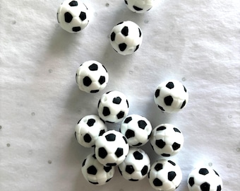 Può includere: Un gruppo di piccole perline rotonde progettate per assomigliare a palloni da calcio. Ogni perlina è bianca con segni neri a forma di pentagono, che imitano il design classico del pallone da calcio. Le perline sono sparse su una superficie chiara e strutturata.
