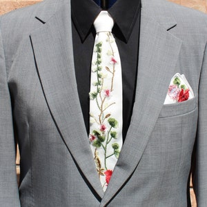 May include: A gray suit jacket with a white tie featuring embroidered flowers and a white pocket square with embroidered flowers.