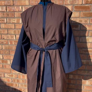 May include: A brown and navy blue robe-style garment with a matching belt tied at the waist. The robe has wide sleeves and a sleeveless brown outer layer. The garment is displayed on a dress form against a brick wall.