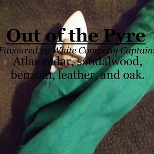 Puede incluir: Un objeto decorativo de madera blanca envuelto en una tela verde. El texto "Out of the Pyre" está escrito en negro sobre el objeto. El texto "Favoured by White Company Captains" está escrito en negro debajo del objeto. El texto "Atlas cedar, sandalwood, benzoin, leather, and oak." está escrito en negro debajo del texto "Favoured by White Company Captains".