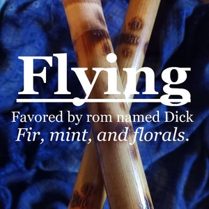 May include: Two brown wooden sticks crossed over each other. The sticks have a dark brown stain and are slightly textured. The text "Flying" is written in white letters above the sticks. The text "Favored by rom named Dick Fir, mint, and florals." is written in white letters below the sticks.