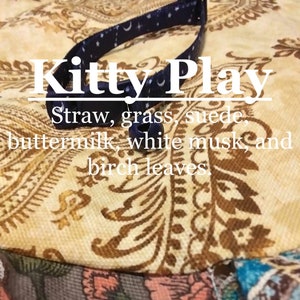 May include: A blue cat collar with white stars and a silver bell. The collar has the text "Kitty Play" printed on it. The text below the collar reads "Straw, grass, suede, buttermilk, white musk, and birch leaves."