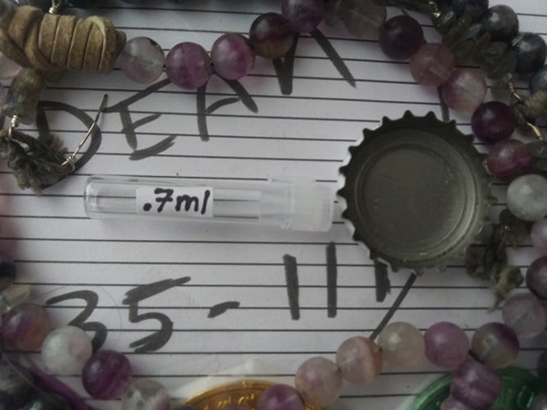 May include: A clear glass bottle with a silver cap and a label that reads ".7ml". The bottle is surrounded by purple and white beads.